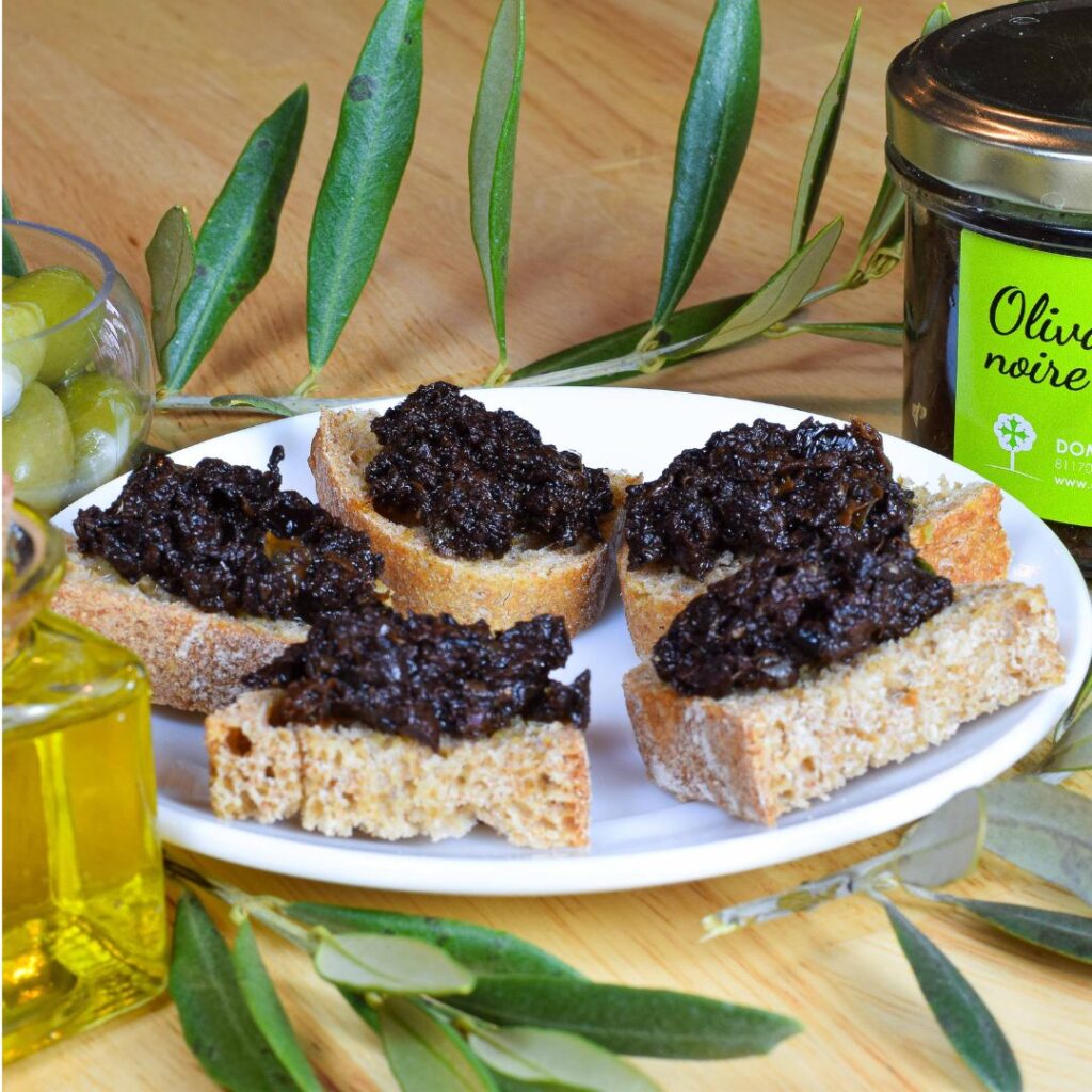 tartine d'olivade noire du domaine rigaud sur une assiette blanche avec un pot olives noires et d'huile d'olive entourée de feuille d'oliviers